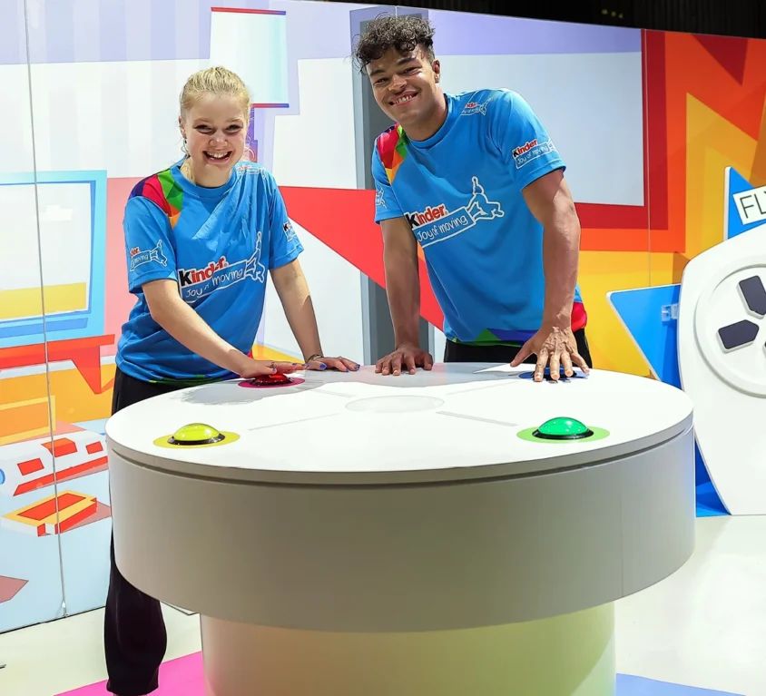 Zwei Markenbotschafter / Influencer lehnen auf einem Spieltisch. Große Arcade-Buttons sind in die Oberfläche integriert um das Rhytmusgefühl der Kinder zu testen.