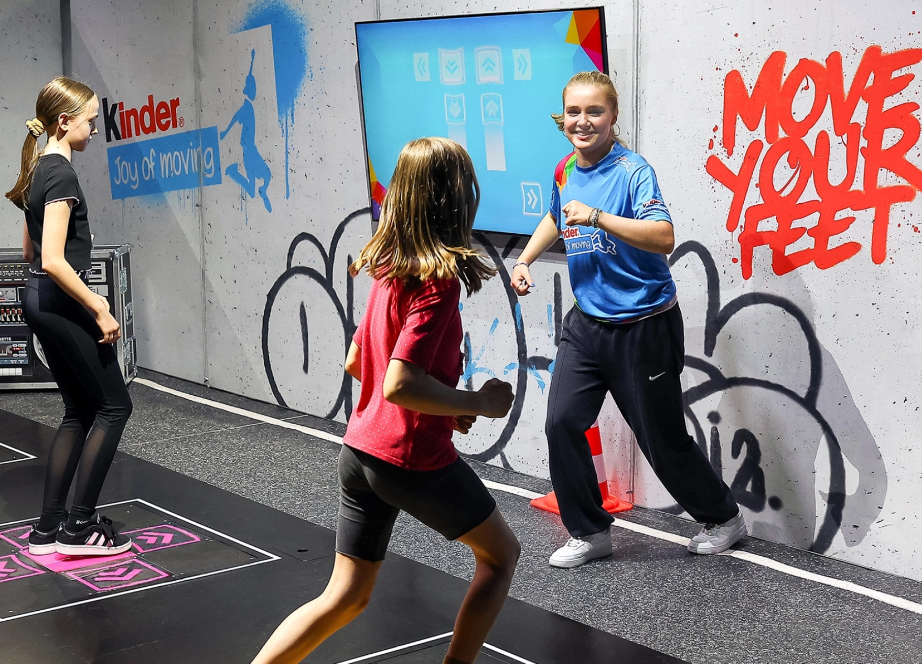 Kinder tanzen zusammen mit einem Influencer auf dem Joy of Moving Event auf Dance Pads. Auf einem Bildschirm werden die Bewegungen der Füße vorgegeben.