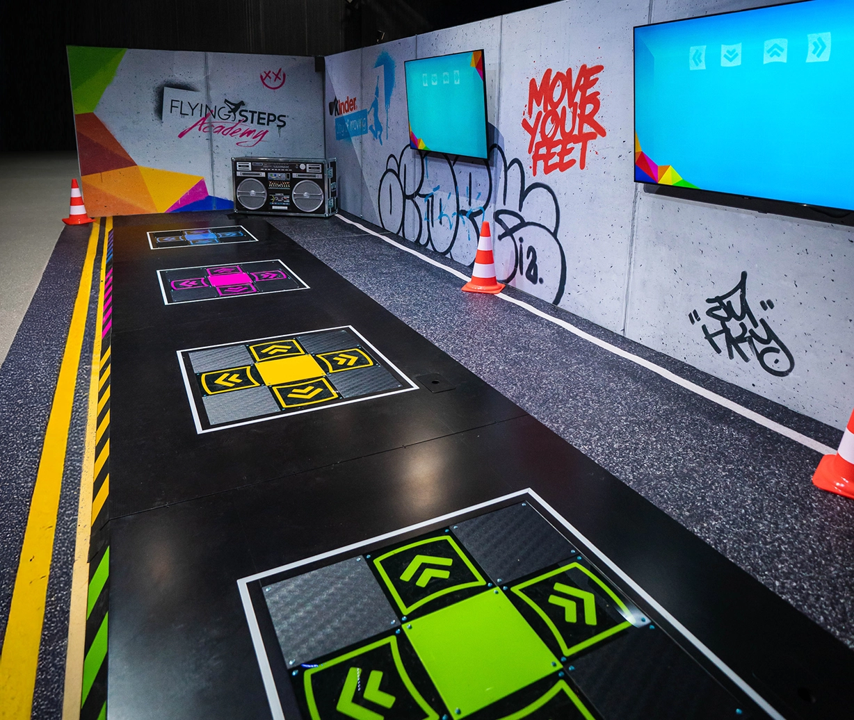 Dance Pads in einer Event-Kulisse. Auf einem Bildschirm werden die Bewegungen der Füße vorgegeben. Eine große Kulisse einer Boombox steht im Hintergrund