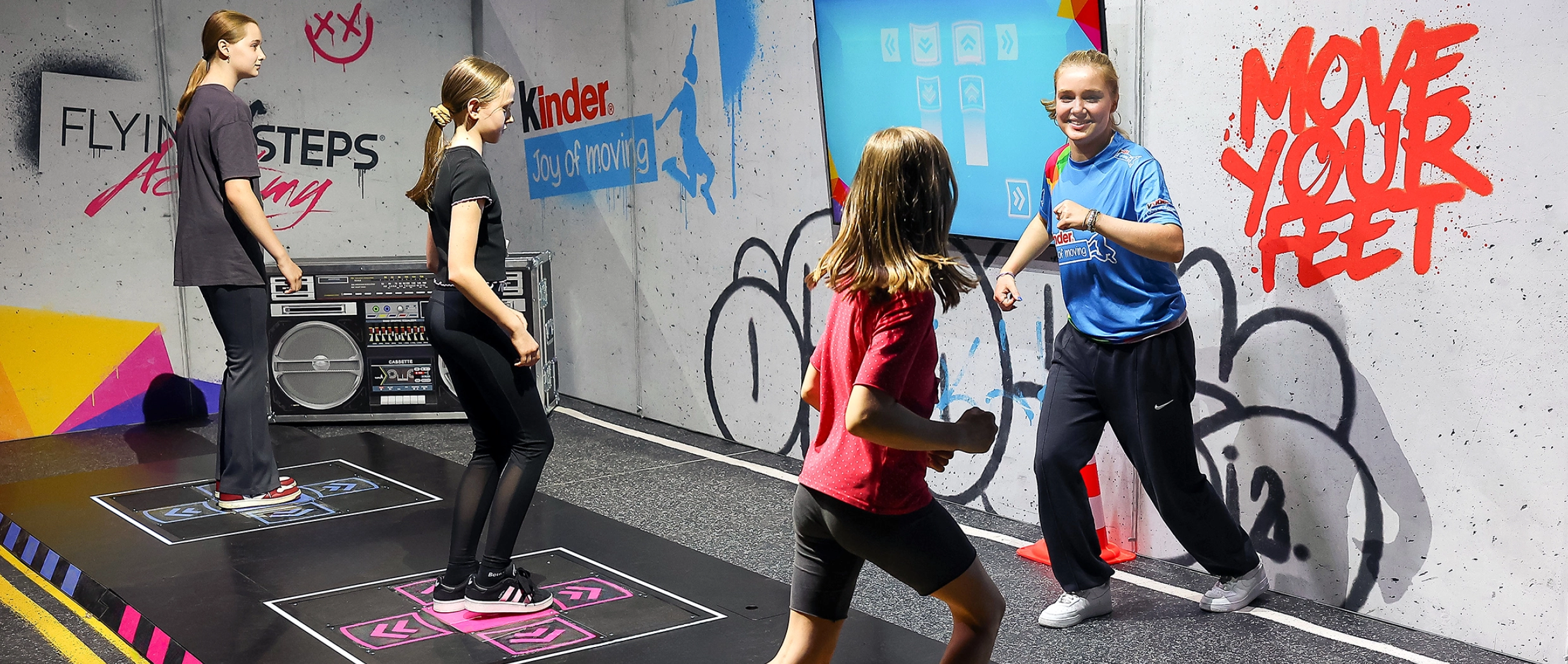 Kinder tanzen zusammen mit einem Influencer auf dem Joy of Moving Event auf Dance Pads. Auf einem Bildschirm werden die Bewegungen der Füße vorgegeben.