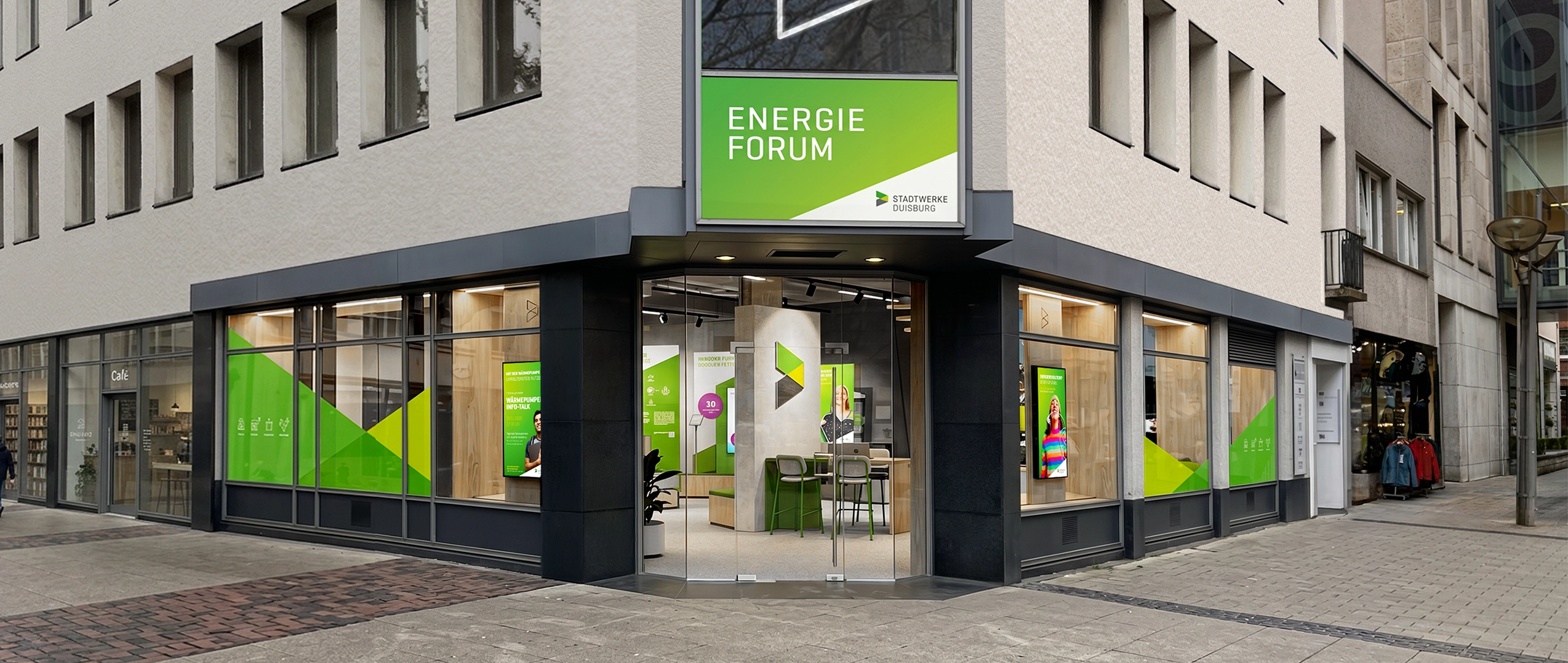 Ehemaliger Retail-Store in Innenstadtlage wird zur multifunktionalen Ausstellungs- und Bürofläche. Der Eckshop mit großer Glastür zeigt die Inneneinrichtung mit Blick auf ein großes Leuchtlogo. In den Schaufenstern sind Holzstelen mit Instore TV Lösung. Die scheiben sind im Stil der Marke foliert.